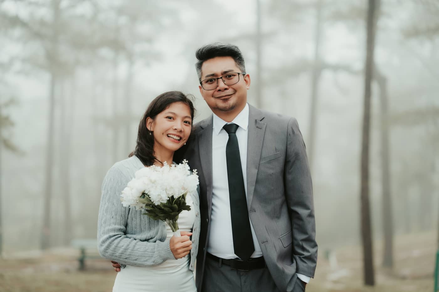 The couple smiling at the camera with a white bouquet in the fog
