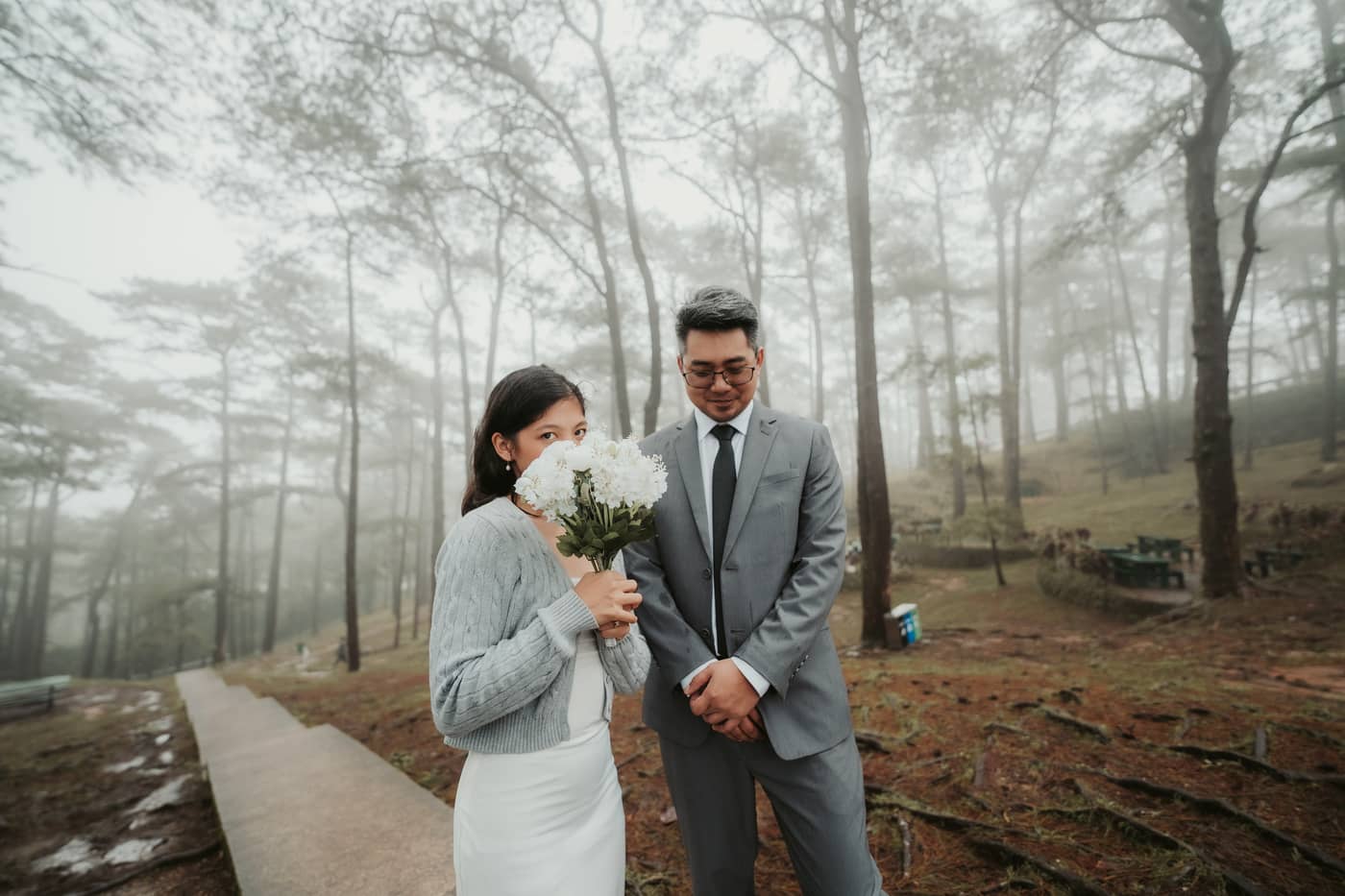 She peeks over a white bouquet while he smiles down, in a misty pine forest