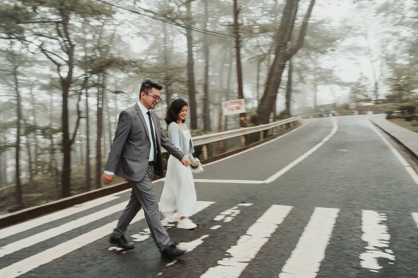 The couple crossing a misty, pine-lined road holding hands